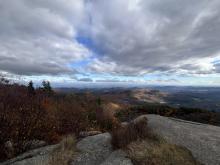 POV from a person on a cliff at the top of a mountain, overlooking a vast forest with a slightly cloudy sky in autumn