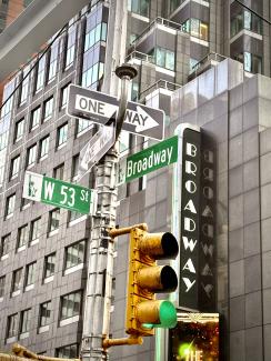 A dynamic evening photo of two Broadway signs in New York City, angled to create an optical illusion where they appear to intersect at 90 degrees. Warm summer dusk tones highlight the vibrant red and white lights against blurred urban buildings.