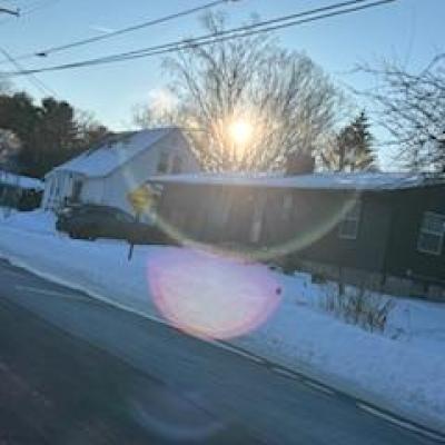 A bluish-tinted, snow-covered street view of a black house with the sun coming in bright through the trees behind the house
