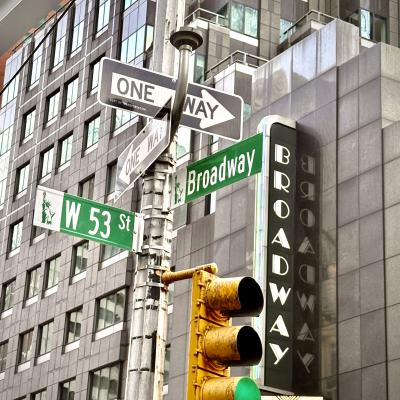 A dynamic evening photo of two Broadway signs in New York City, angled to create an optical illusion where they appear to intersect at 90 degrees. Warm summer dusk tones highlight the vibrant red and white lights against blurred urban buildings.