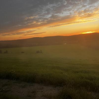 A grassy hill with a background of a sparkling sun of oranges and yellows with blue gray, and white clouds with  silhouettes of hills in the distance.