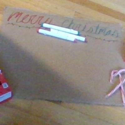A piece of cardboard, on which lays some markers and a box of candy canes, written are the words Merry Christmas